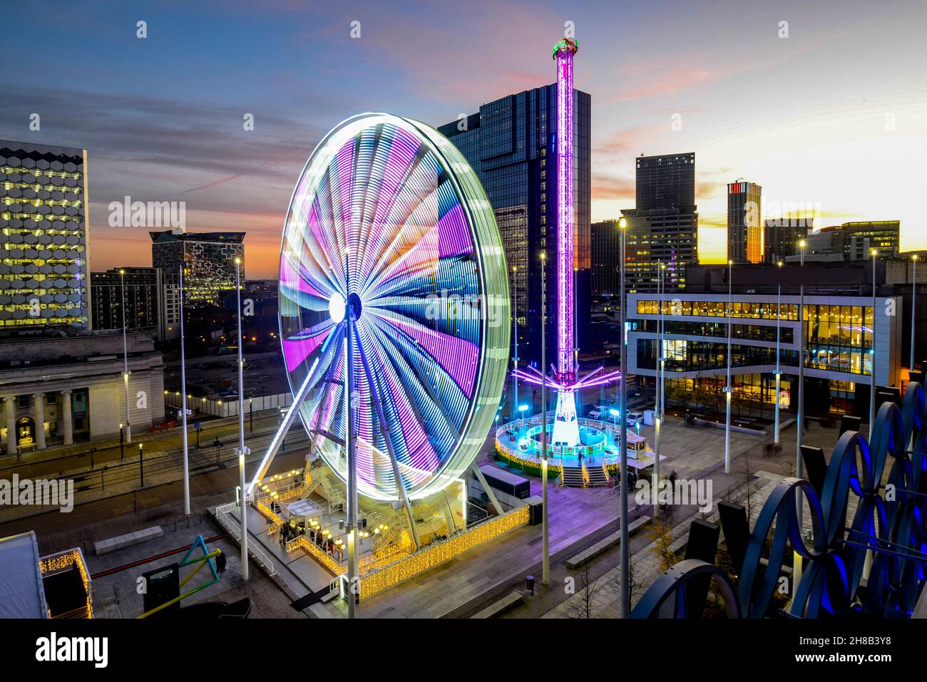 Giostre illuminate nella zona fieristica di Natale, ruota panoramica, e edifici moderni circostanti del centro della città, illuminati al tramonto, dopo il tramonto su una bella, limpida, Foto Stock