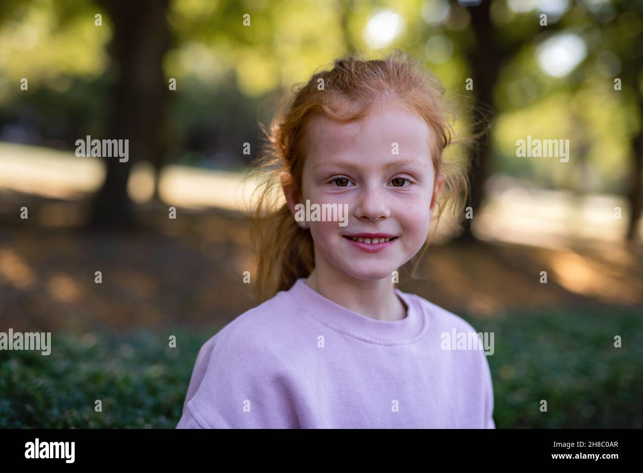 Ragazza piccola felice con frickles e capelli rossi guardando la macchina fotografica e sorridendo all'aperto nel parco. Foto Stock