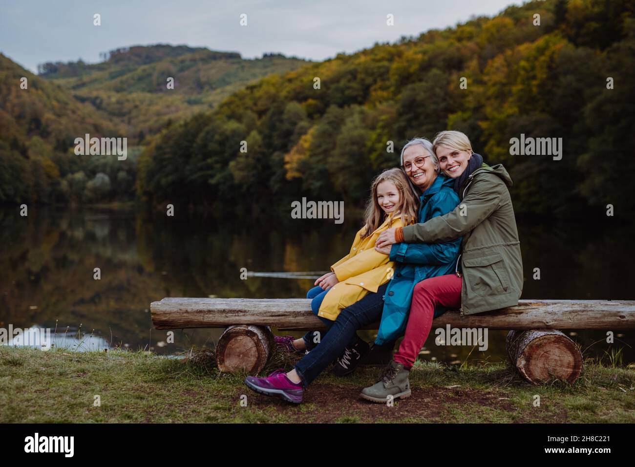 Bambina con madre e nonna seduta su panca e guardando la macchina fotografica outoors vicino al lago. Foto Stock