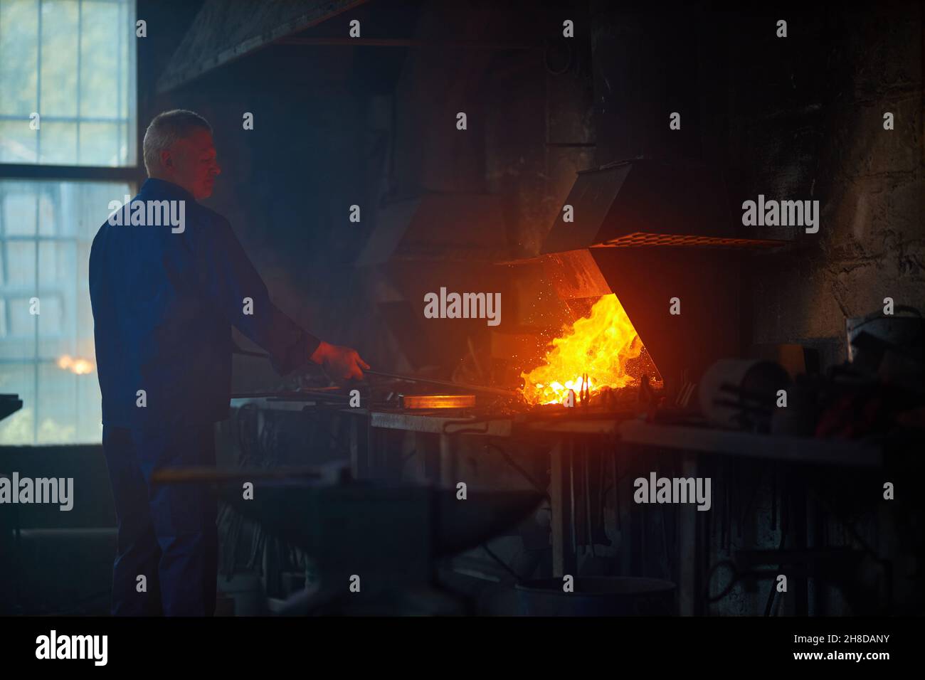 Vista laterale del lavoratore in uniforme blu scuro che fa cancelli in smithy vicino attrezzatura speciale di riscaldamento. Concetto di processo produrre recinzione moderna in garage. Foto Stock