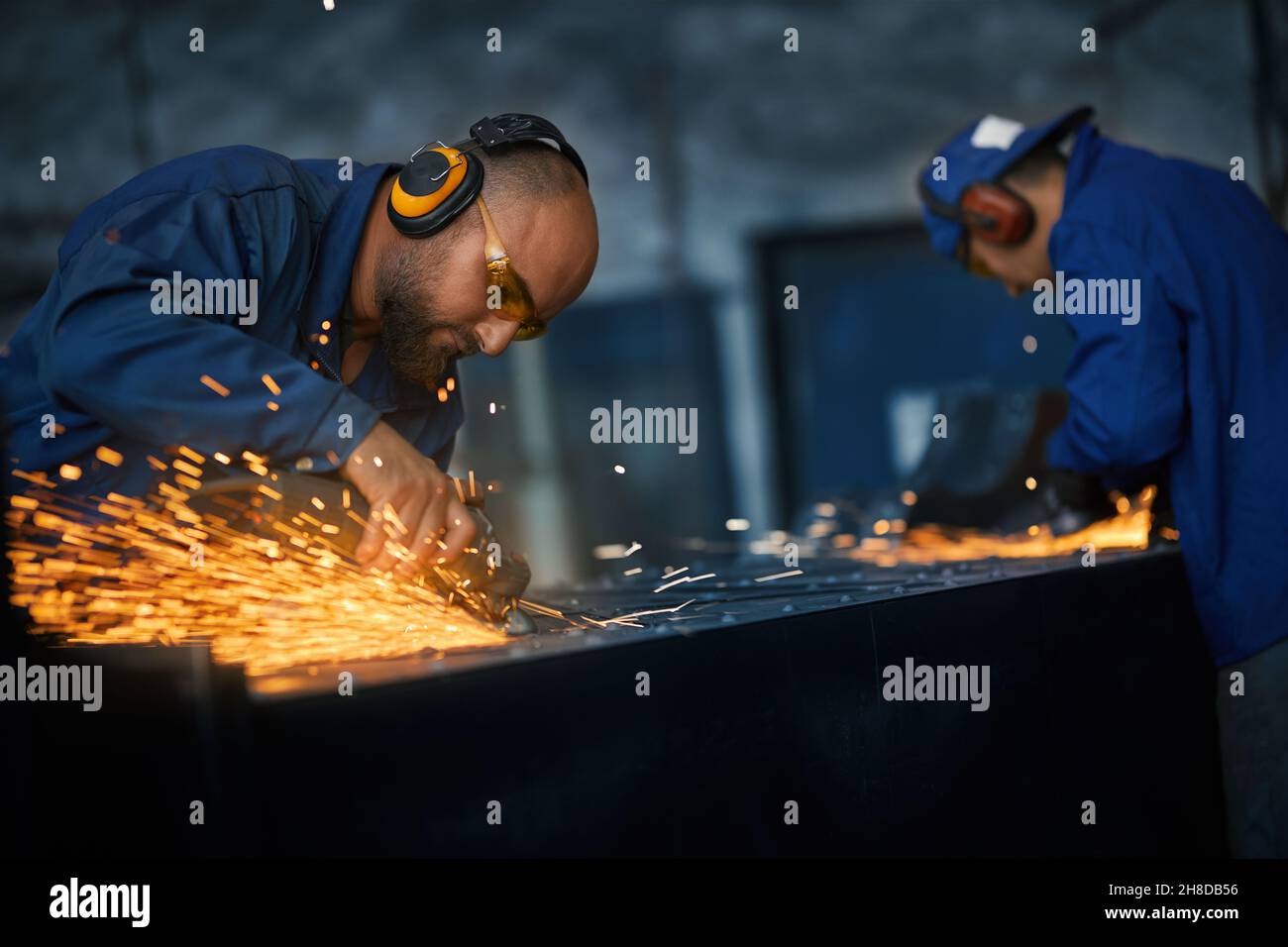 Vista laterale di due giovani lavoratori in speciale blu scuro uniforme e cuffie lucidare o creare metallo con attrezzature speciali vicino alle scintille. Concetto di processo che fa recinto. Foto Stock