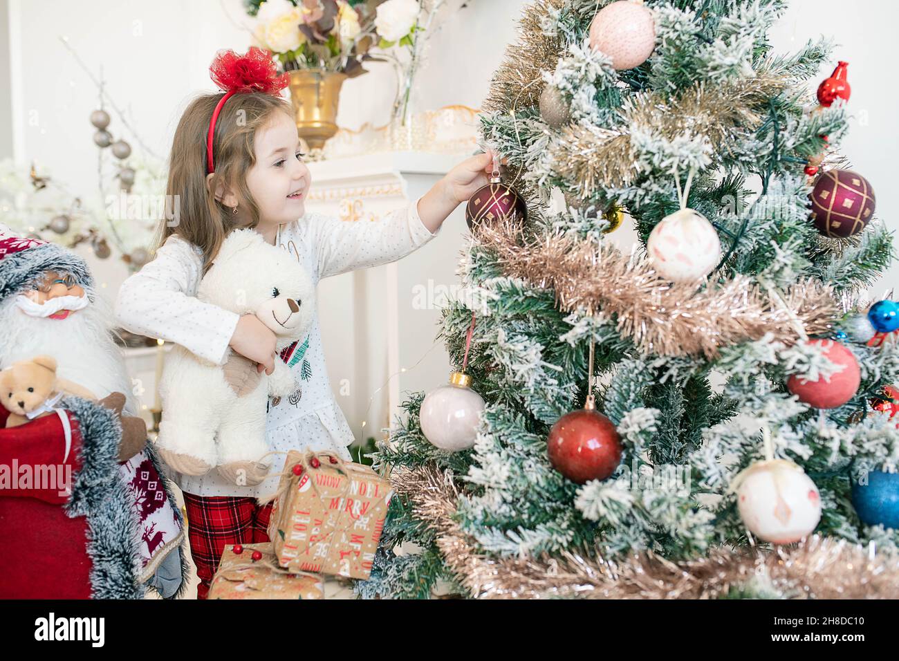 Bella bambina caucasica che indossa pigiami festosi e che tiene in mano un grazioso orsacchiotto mentre aggiunge globi e ornamenti ad un albero di Natale Foto Stock