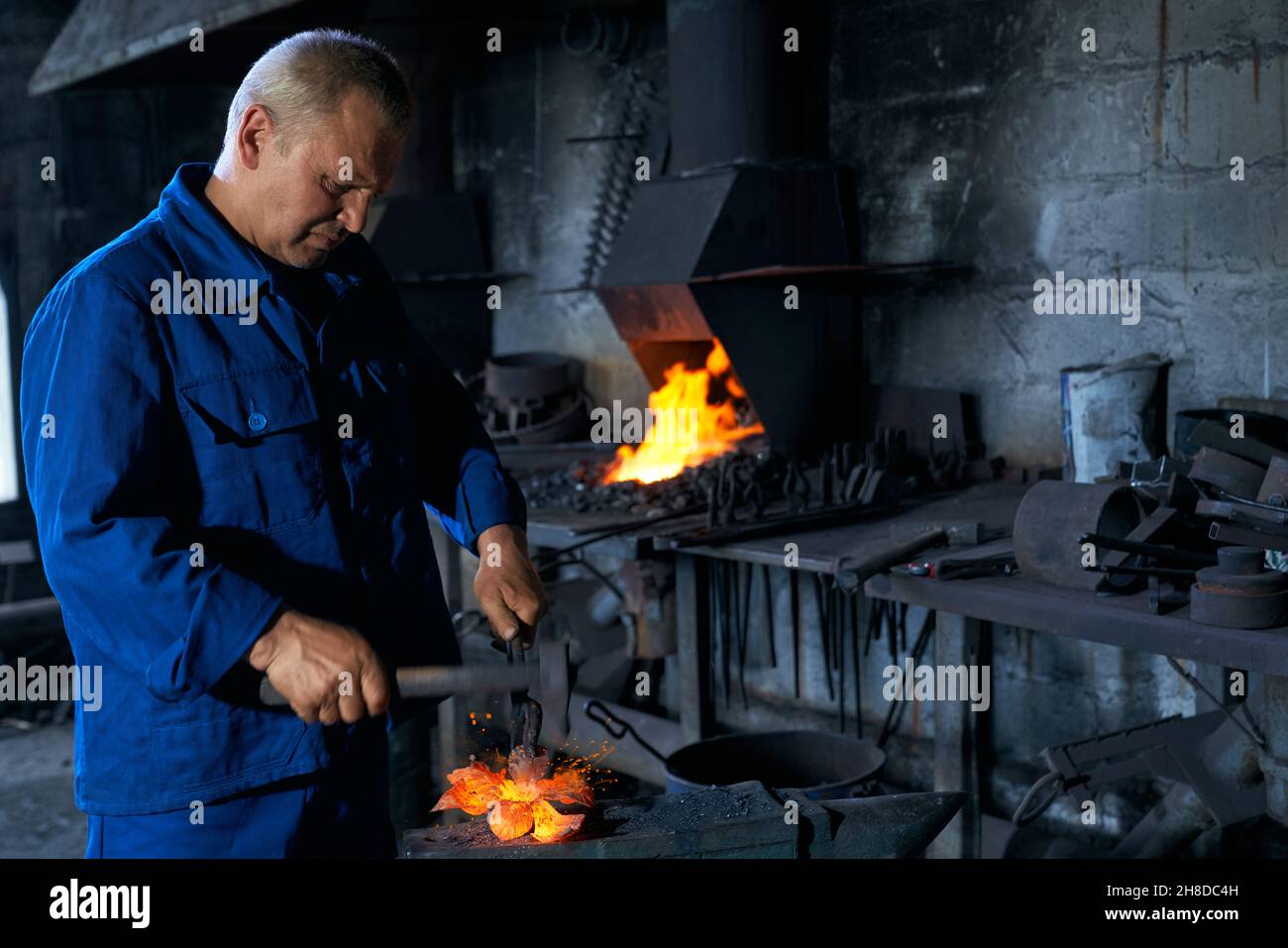 Vista laterale di un uomo anziano in speciale uniforme blu scuro lavorando con attrezzi speciali e creando attrezzature in metallo vicino al riscaldamento. Concetto di processo che crea recinzione in smithy. Foto Stock
