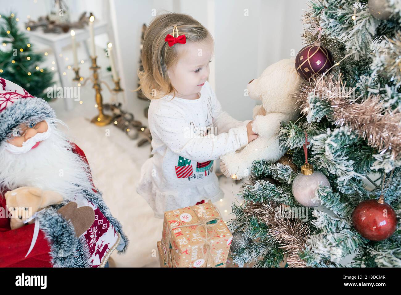 Ragazza caucasica carina indossando pigiami festosi e giocando con il suo orsacchiotto vicino ad un albero di Natale decorato con grandi globi e neve artificiale Foto Stock