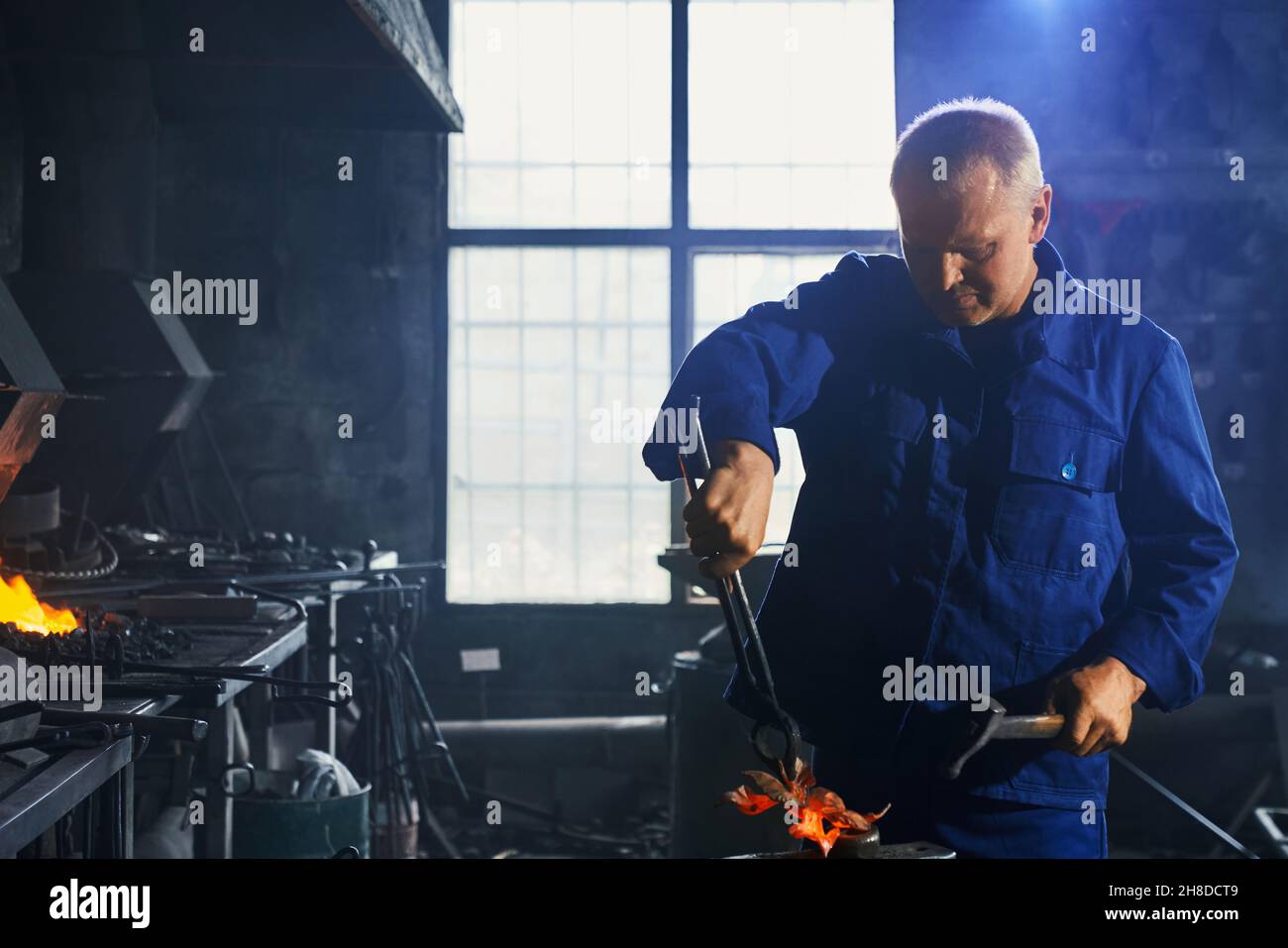 Vista frontale dell'uomo adulto in uniforme blu scuro vicino al riscaldamento attrezzatura che lavora con martello e pinze per creare decorazioni per recinzione. Concetto di uomo che crea decorazioni in smithy. Foto Stock