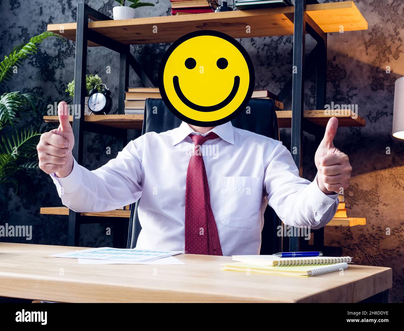 Un uomo d'affari gioioso al tavolo con un volto sorridente invece di una testa. Foto Stock