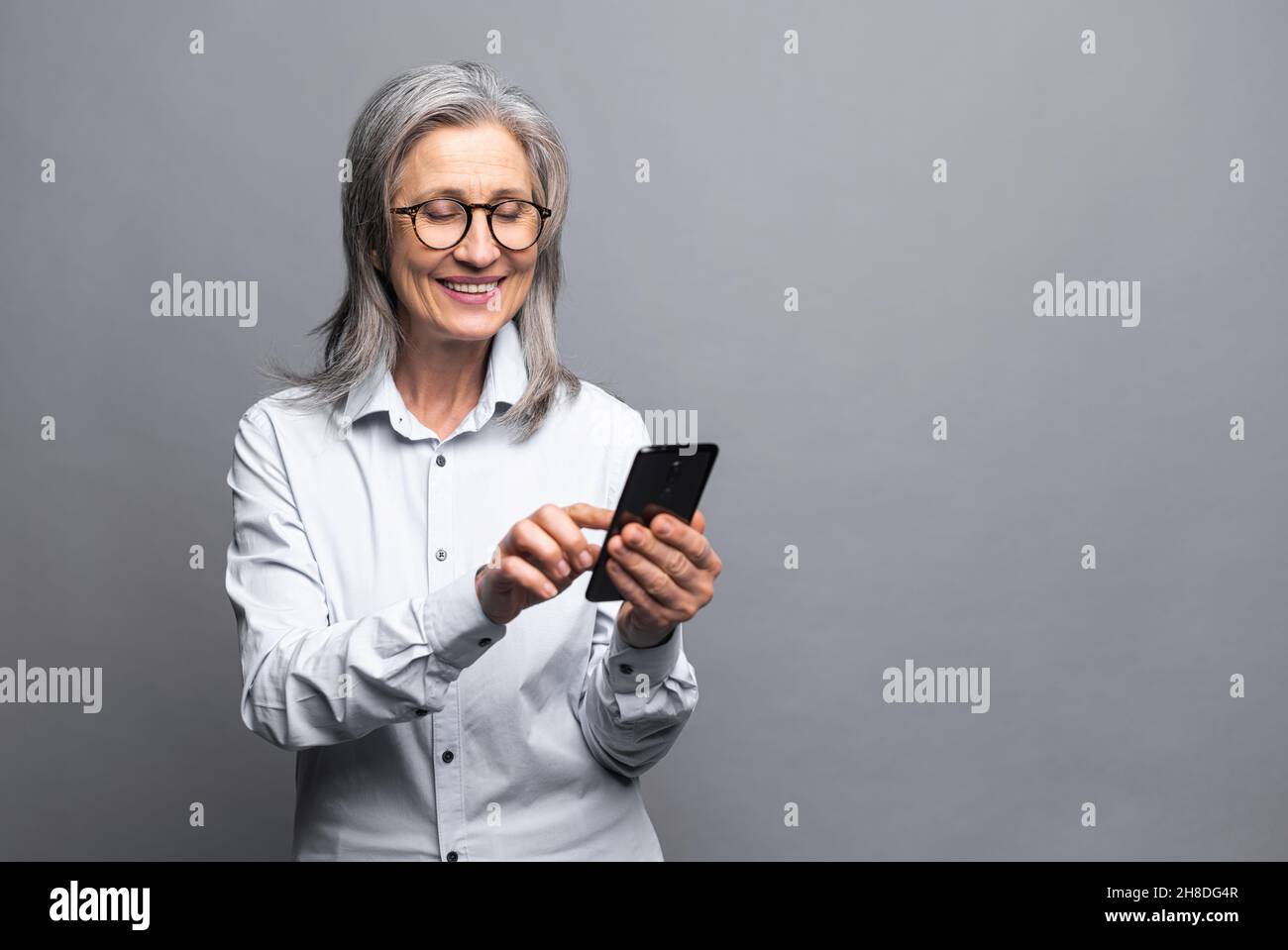 Allegro positivo felice anziano donna d'affari grigio-pelata usando smartphone in piedi isolato su grigio, donna matura in abbigliamento formale tocco touchscreen, scorrimento news feed, messaggi, SMS Foto Stock