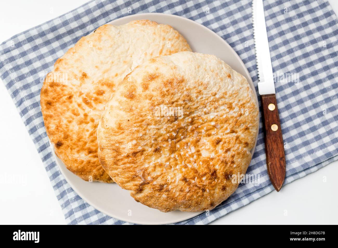 Lafs del pane rotondo piatto del grano in un piatto, con un coltello e un panno blu della cucina sopra il tavolo bianco Foto Stock