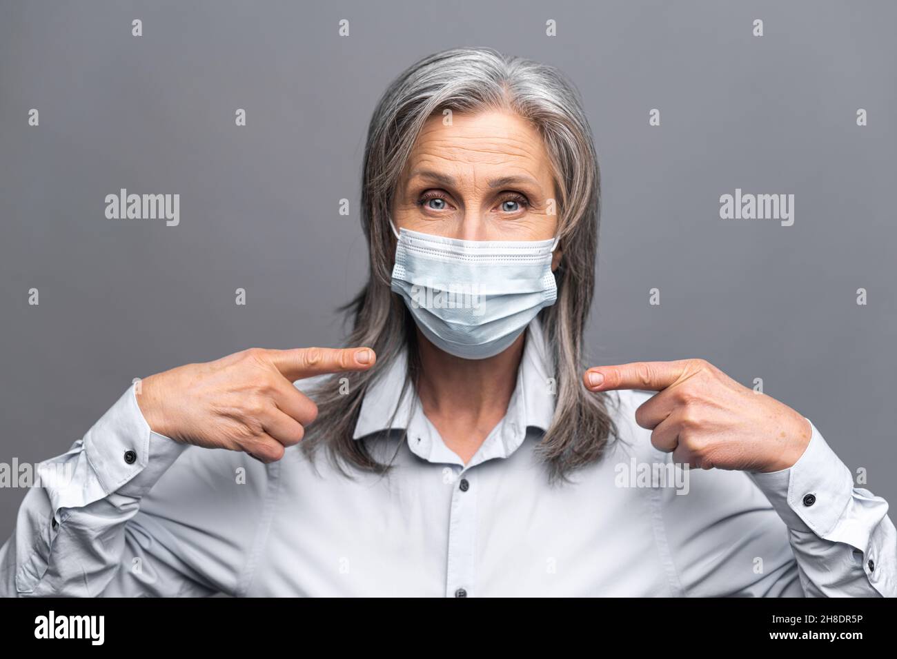 Donna anziana seria in abbigliamento formale e maschera medica sui punti del viso dita isolate su grigio, ritratto close-up di dipendente ufficio femminile proteggendosi da malattie virali, raccomanda misure Foto Stock