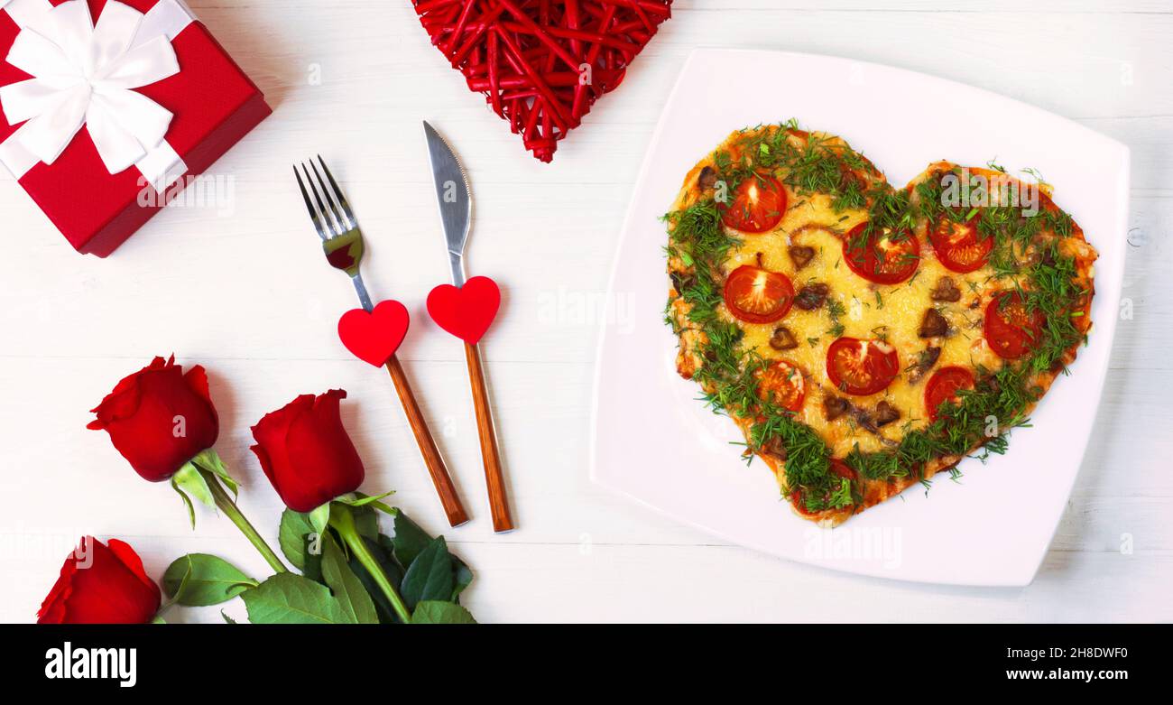 San Valentino cibo. Pizza a forma di cuore con verdure, su sfondo bianco di legno. Rose rosse accanto a un regalo in una scatola rossa. Posate con cuori. Vista superiore Foto Stock