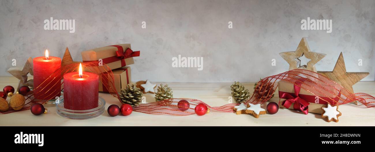 Decorazioni natalizie in formato panoramico con candele rosse, baubles, nastro, stelle in legno e regali avvolti in natura su un tavolo, parete grigio chiaro con co Foto Stock