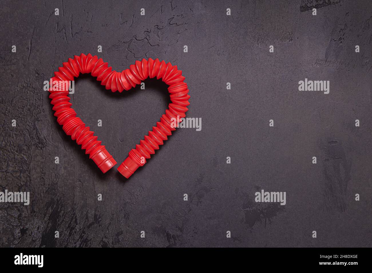 Rosso brillante steso pop it tubo bambini giocattolo piegato a forma di cuore. Giocattolo antistress in plastica su sfondo nero in pietra testurizzata. Disposizione piatta. Copia di grandi dimensioni Foto Stock