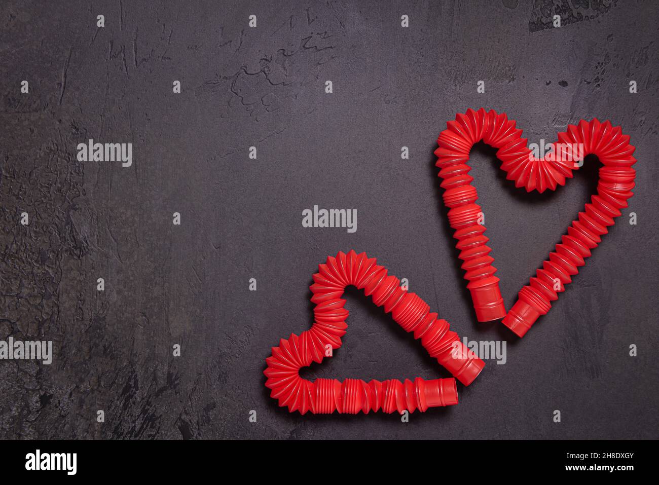 Due stese rosso pop tubo per bambini giocattoli piegati a forma di cuori. Giocattoli antistress in plastica su sfondo di pietra nero testurizzato. Disposizione piatta. Copia Foto Stock