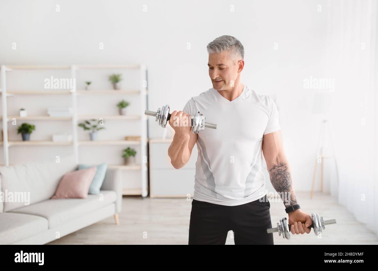 Concetto di formazione nazionale. Uomo maturo sportivo che si esercita con i manubri a casa, allenando il suo bicipite, spazio libero Foto Stock