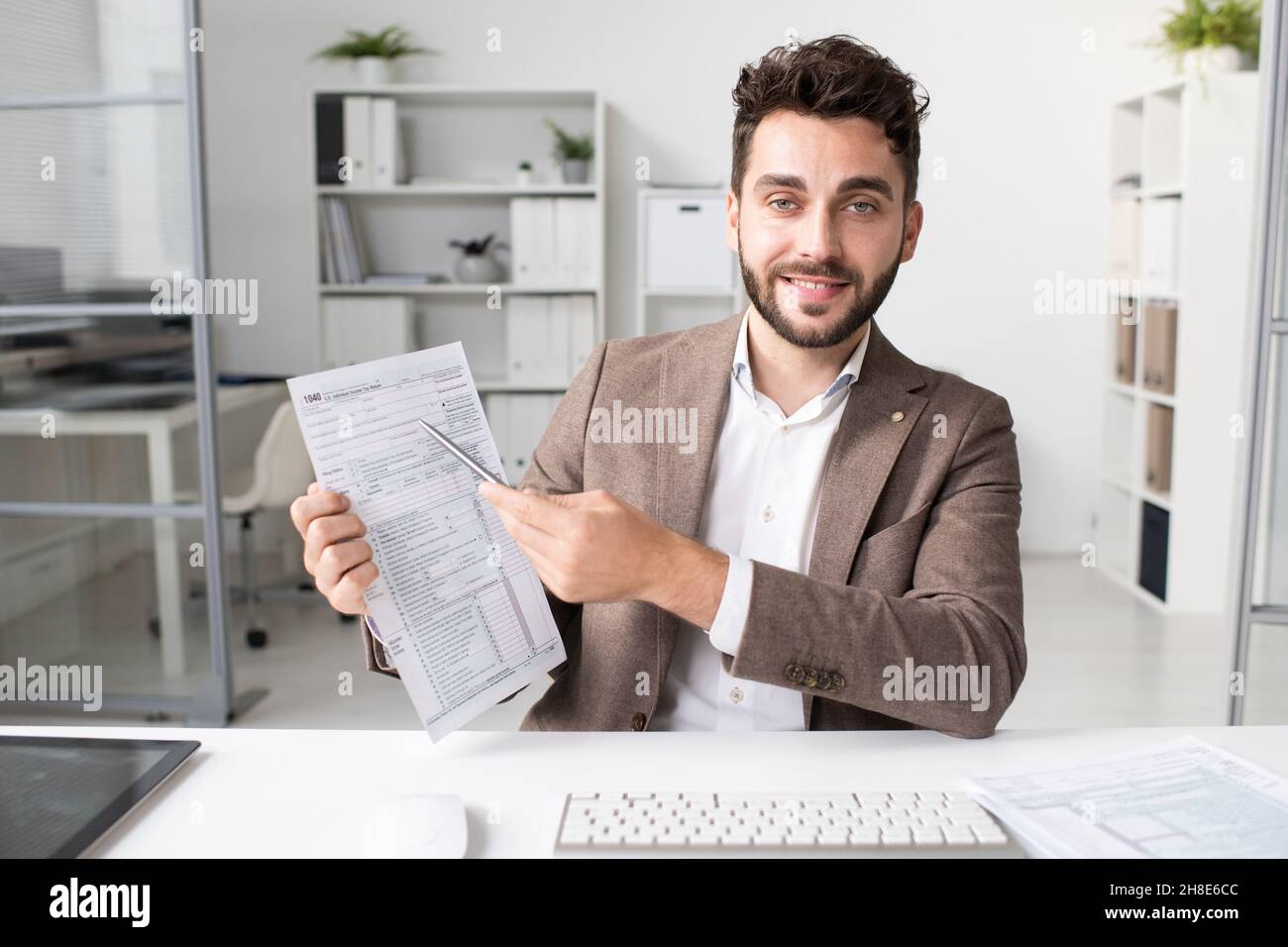 Ritratto di ragioniere sorridente barbuto in giacca seduta alla scrivania in ufficio moderno e indicando la forma di imposta Foto Stock