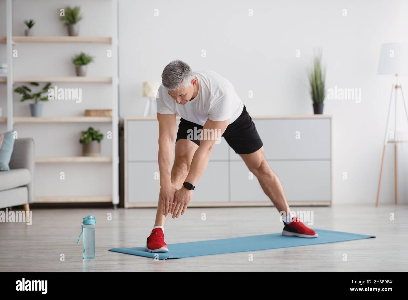 Uomo anziano sportivo che allunga le gambe, esercitandosi a casa, facendo esercizi di flessibilità e conducendo stile di vita sano, a tutta lunghezza Foto Stock