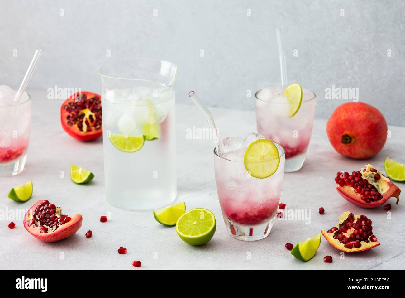 Rinfrescante bevanda di melograno in bicchieri con semi di melograno e ghiaccio, cannucce di calce e vetro, brocca di acqua fredda con agrumi, melograni tagliati Foto Stock