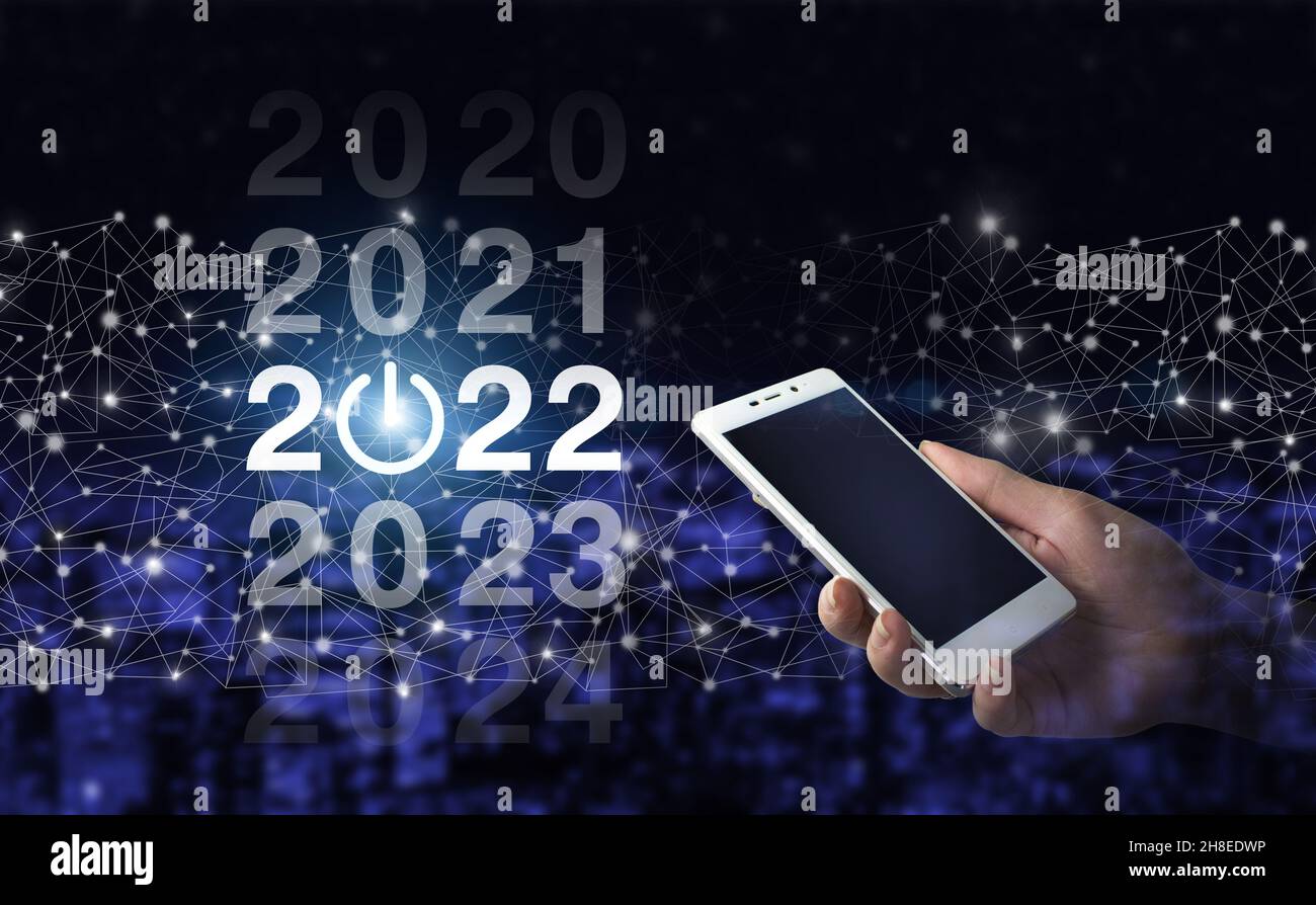 Un uomo d'affari benvenuta anno 2022. Smartphone bianco con hologram digitale 2022 sullo sfondo sfocato scuro della città. Anno di carico dal 2021 al 2022. Foto Stock