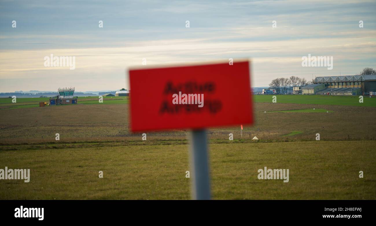 Focus dietro il luminoso rosso airfield attivo segno pista sugli edifici a campi d'aria e oltre la pista di erba, Wiltshire UK Foto Stock