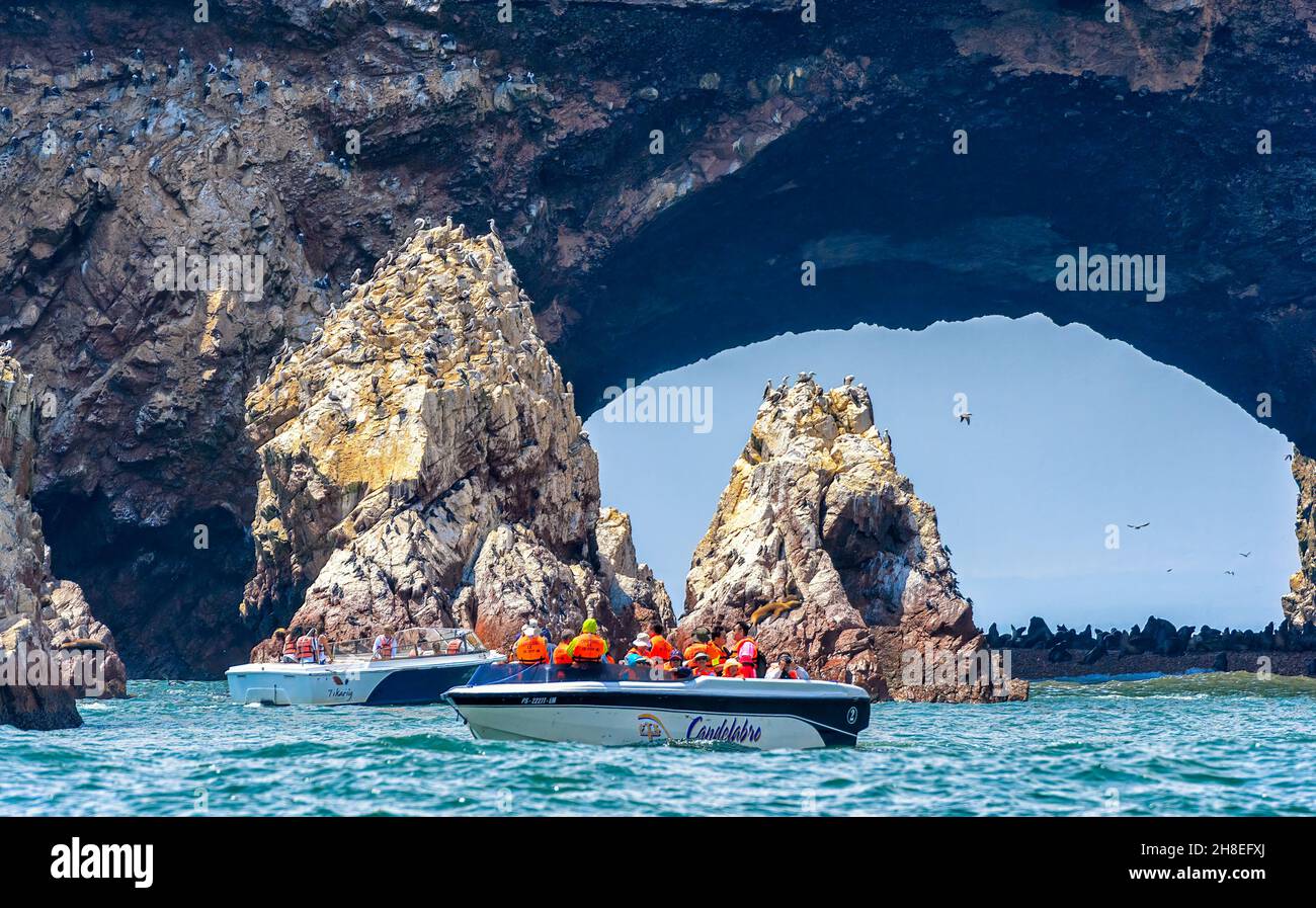 I turisti, in barca, guardano gli uccelli che volano sotto l'arco di roccia naturale nella Riserva Naturale delle Isole Ballestas al largo della costa di Paracas in Perù Foto Stock