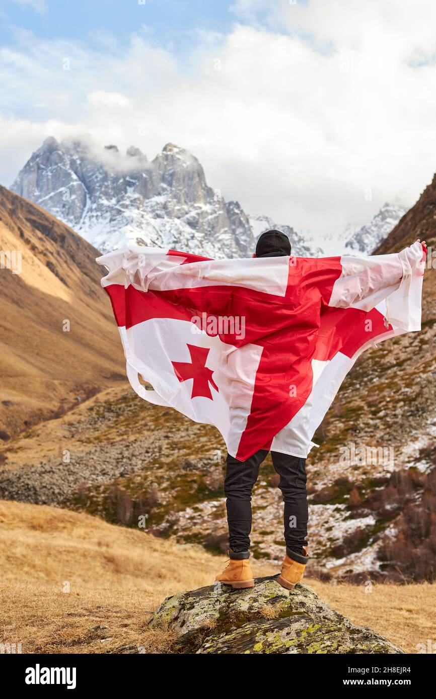 La vista posteriore del giovane felice in posa con la bandiera nazionale della Georgia in piedi all'aperto guarda le vette innevate Foto Stock