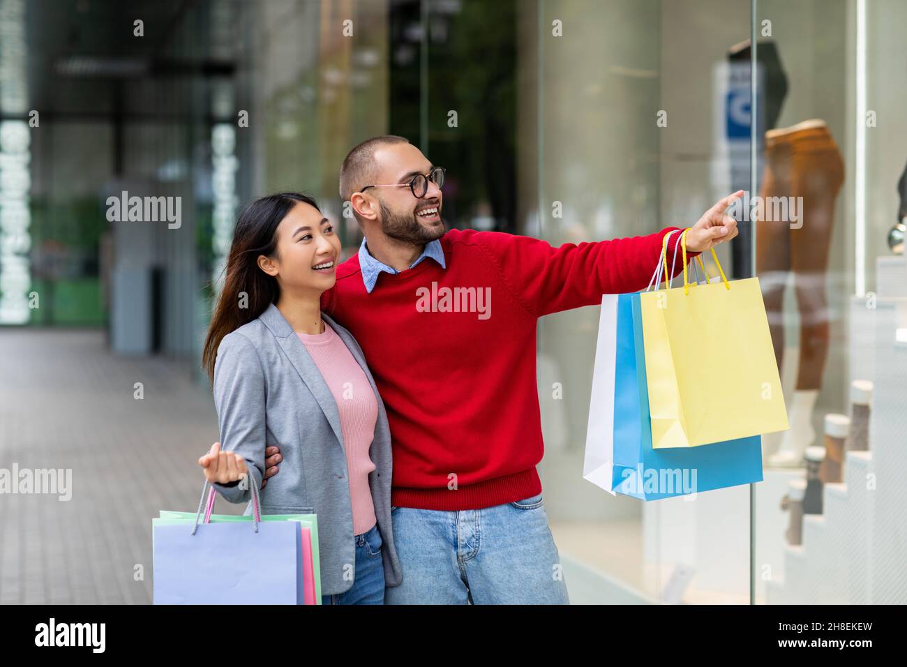 Giovane coppia interrazziale affettuosa in piedi sulla strada con borse shopping, che punta alla vetrina vicino al centro commerciale della città Foto Stock