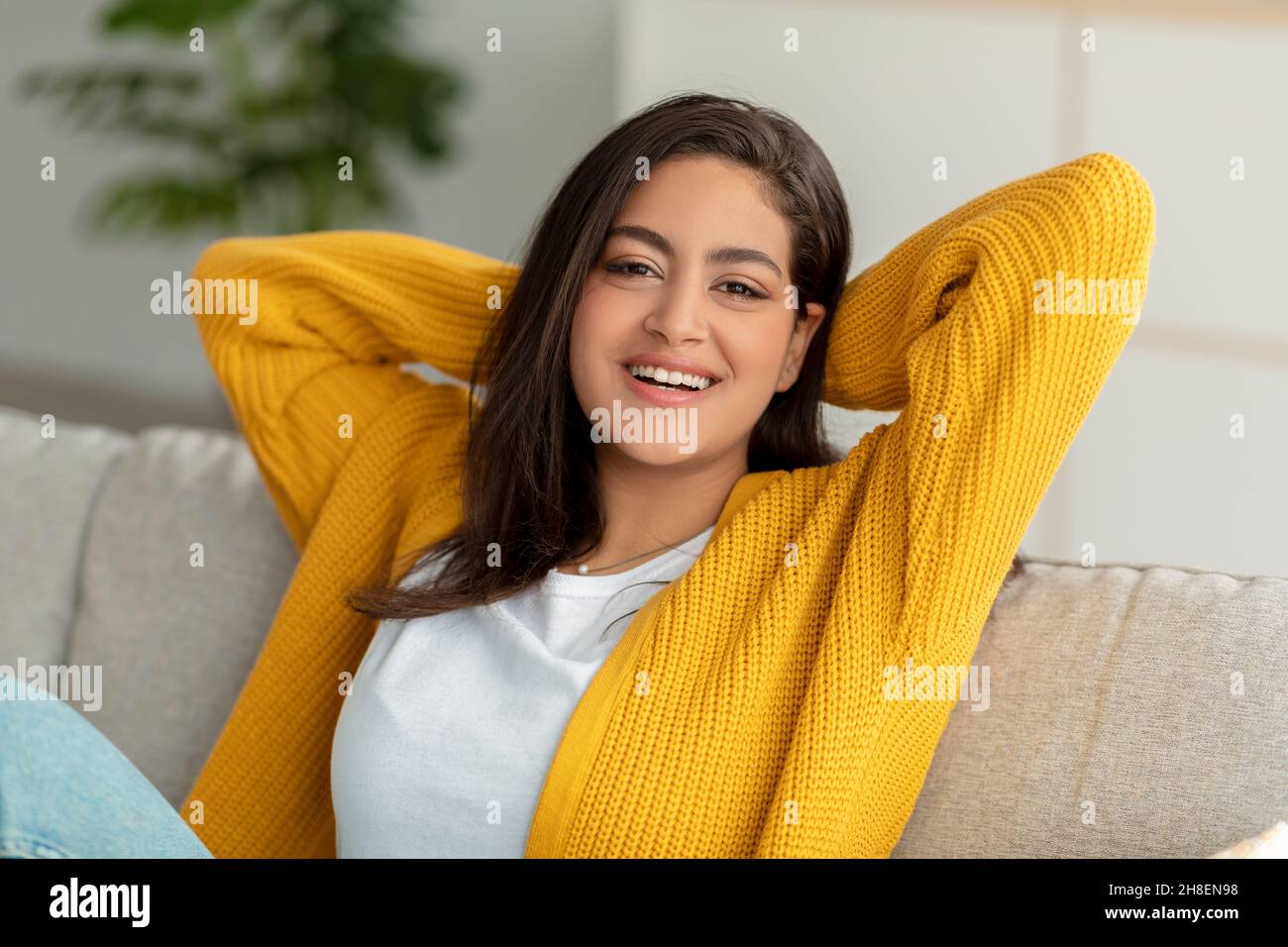 Riposatevi e rilassatevi a casa vostra. Una donna musulmana felice gettò le mani dietro la testa e sorrise alla macchina fotografica, seduto sul divano Foto Stock