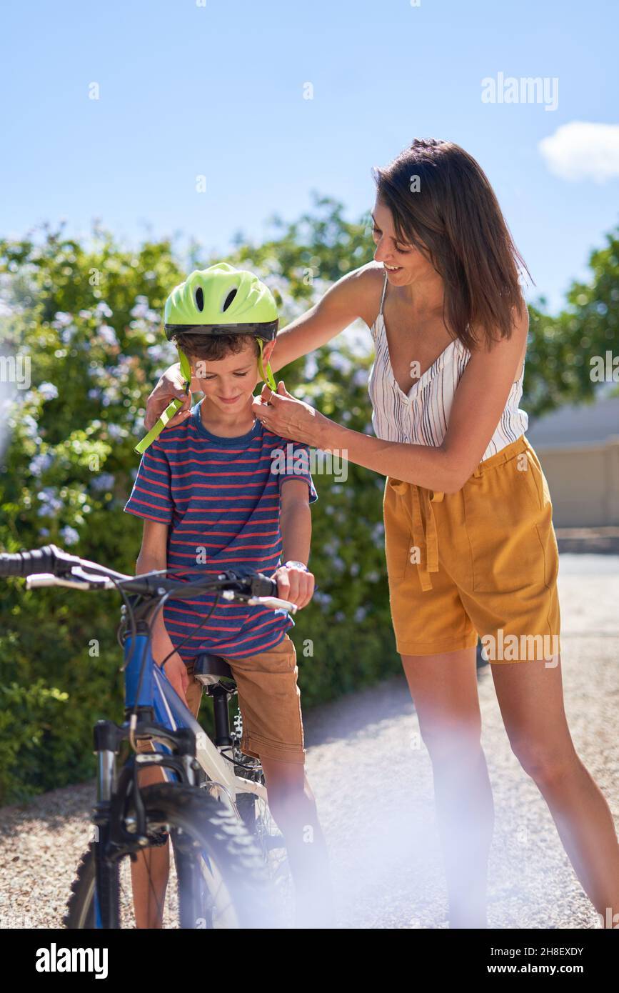 Madre che aiuta il figlio con casco in bici in strada soleggiata Foto Stock