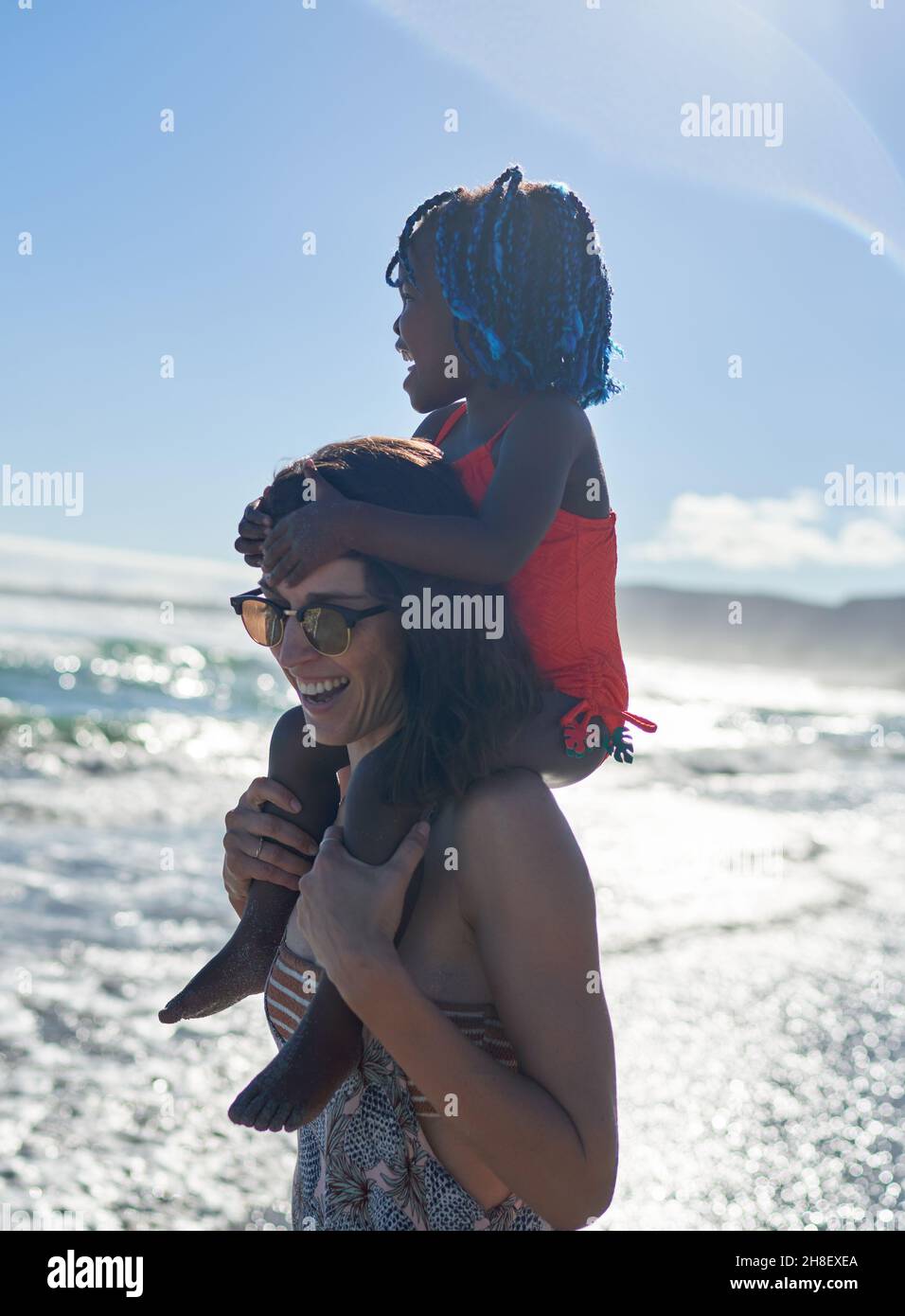 Felice madre che porta la figlia carina sulle spalle sulla spiaggia soleggiata dell'oceano Foto Stock
