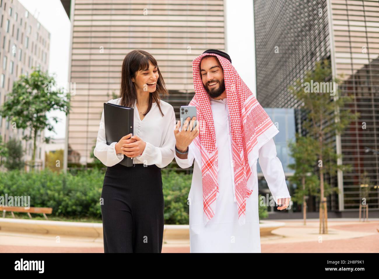 Felice barbuto arabo cliente maschile in abiti tradizionali mostrando cellulare al manager femminile ottimista mentre si cammina per strada contro grattacieli durante il business meeting in centro Foto Stock