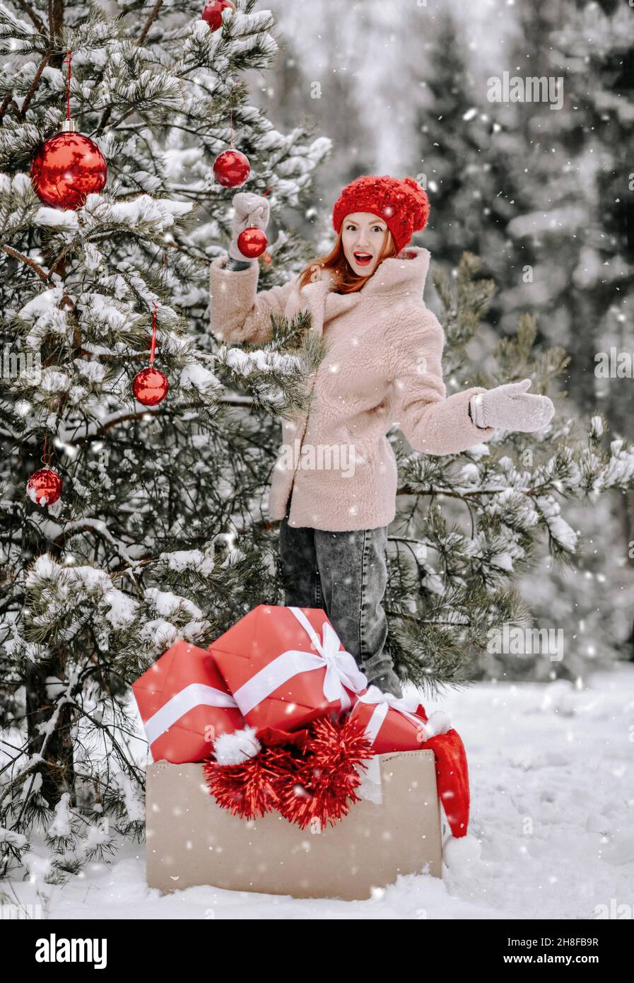 Giovane donna positiva sorpresa in abbigliamento elegante in piedi vicino albero di Natale decorato con mucchi di scatole presenti all'aperto Foto Stock