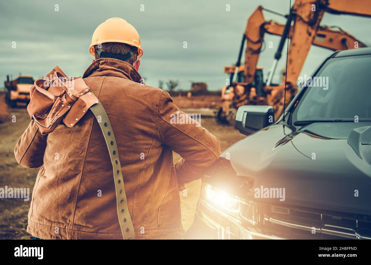 Lavoratore appaltatore con cintura portautensili sulla spalla accanto a camion di raccolta, escavatori e altre attrezzature pesanti in background. Tema industriale. Foto Stock