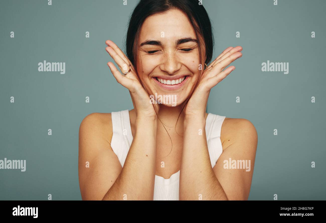 Giovane donna eccitata sorridendo con gli occhi chiusi in uno studio. Giovane donna felice abbracciando il suo volto mentre si alza da sola contro uno sfondo studio. Foto Stock