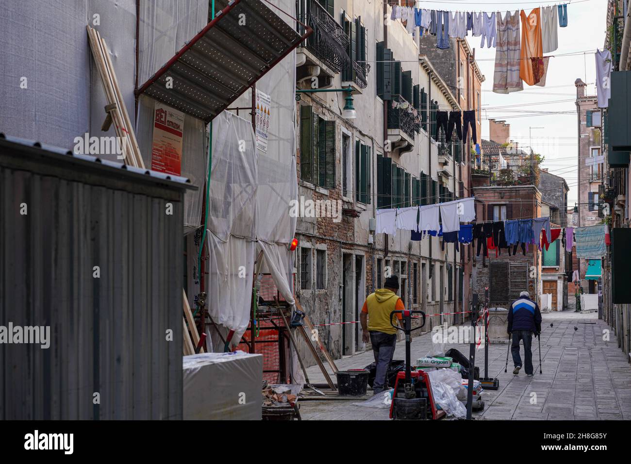 Vista su un piccolo vicolo di Venezia attraverso il quale un uomo con due stampelle cammina. Sopra di lui sospese il lavaggio. In primo piano un operaio di costruzione. Foto Stock