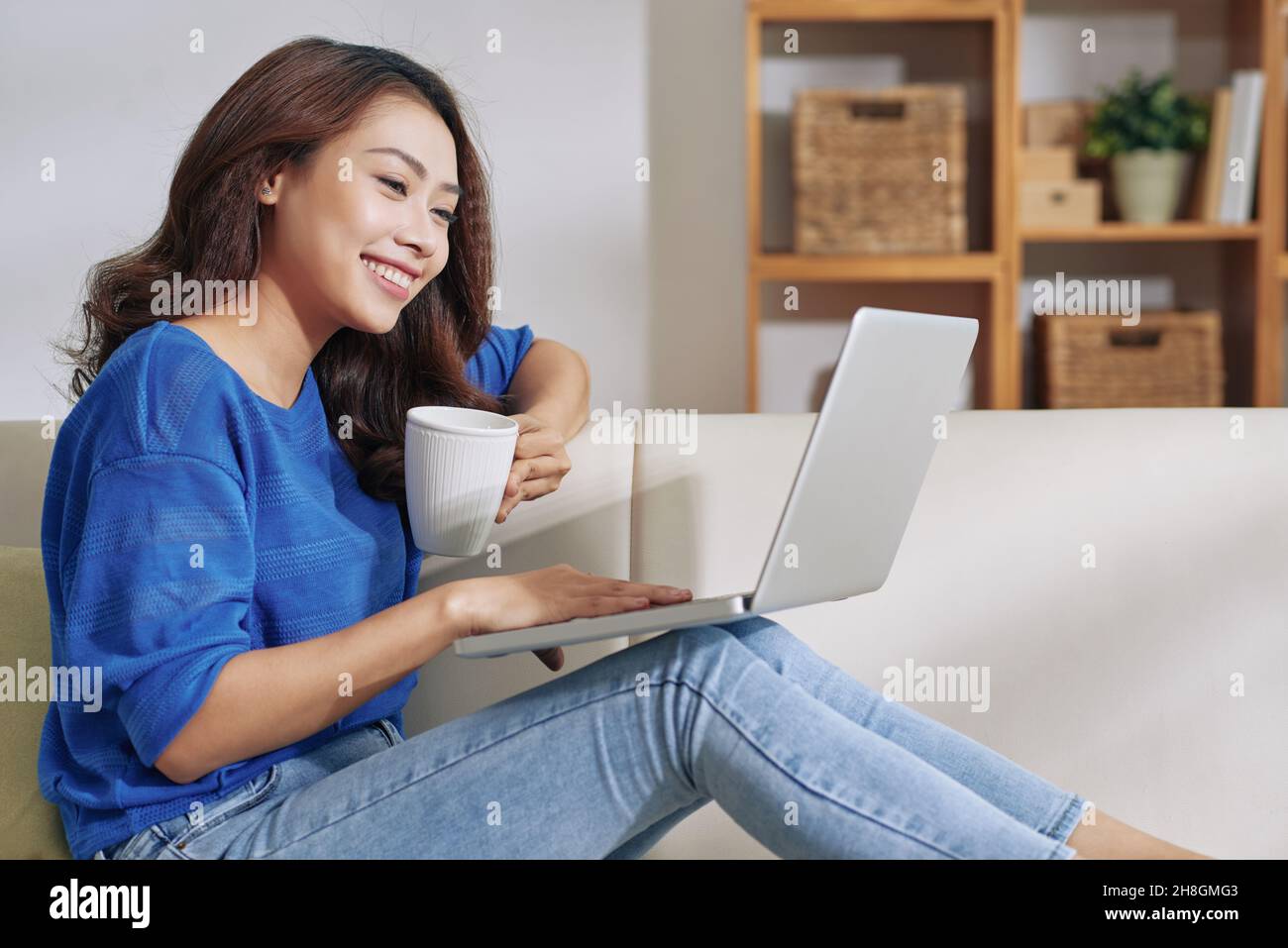 Sorridente giovane donna asiatica seduta con un computer portatile in ginocchio e bere caffè mentre si lavora a casa Foto Stock