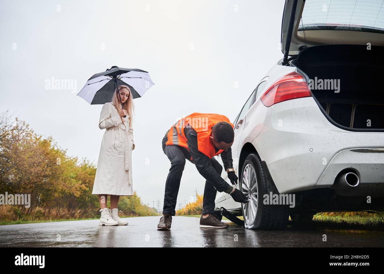 Maschio meccanico auto svitando dadi di aggetto su ruota auto mentre donna elegante tenendo ombrello. Giovane uomo in gilet riparazione donna automobile sulla strada. Concetto di servizio stradale di emergenza. Foto Stock