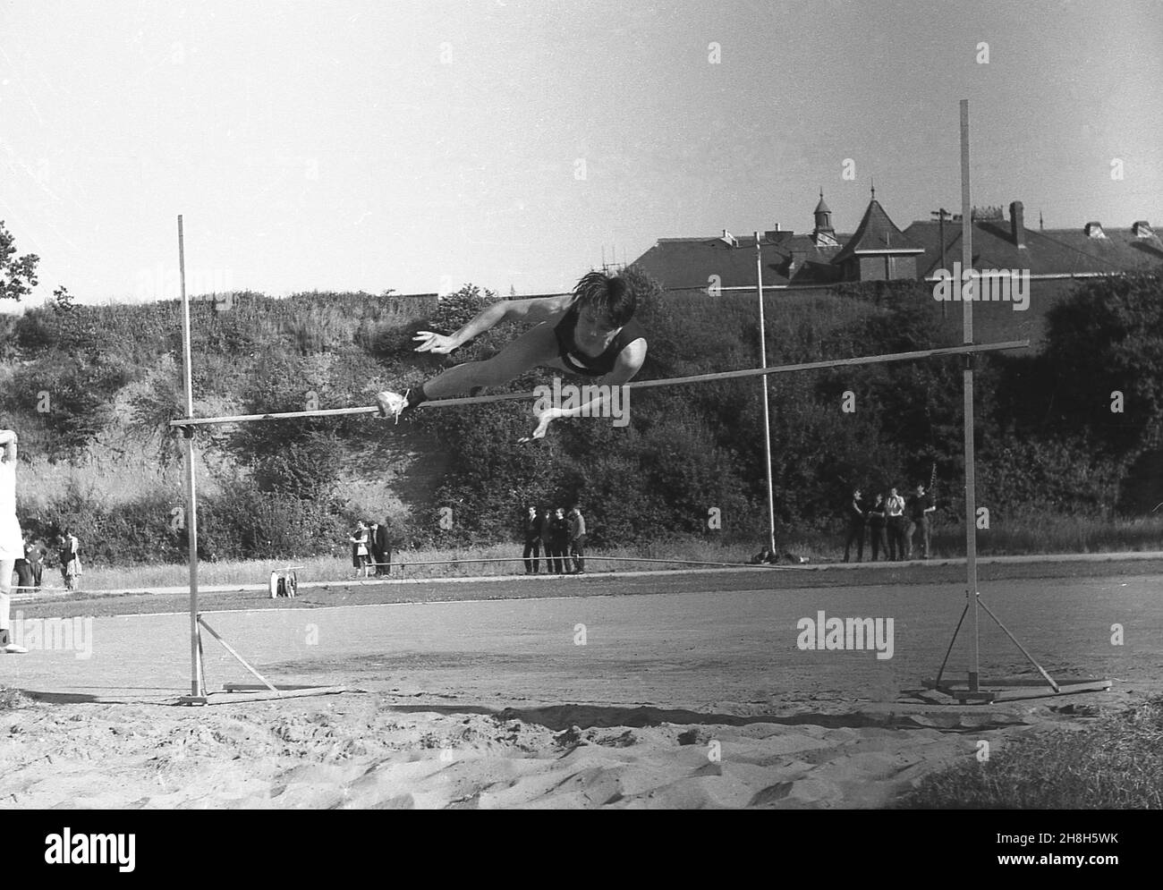 1964, storico, sport scolastici, ora legale, Devon, Inghilterra, Regno Unito e fuori accanto ad un tradizionale pozzo di sabbia, un giovane che affronta il salto in alto, un evento pista & campo. Tecnica interessante, assomiglia ad una forma del Western Roll, dove la gamba di piombo è calciata in su sollevando il corpo che ruota sopra la barra con la gamba di coda piegata sotto il piombo. Il Western Roll alla fine sostituì il salto con le forbici originali, di cui c'erano molte varianti diverse, dove il concorrente sbarcò generalmente sui loro piedi. Foto Stock