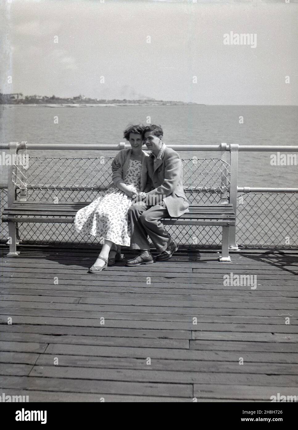 anni '50, storico, uomo e donna seduti a stretto contatto su una panchina su un molo di mare in legno. Alla fine degli anni '20 e indossando i vestiti dell'epoca, sono rilassati, tenendo le mani, magari corteggiando e/o in vacanza, Inghilterra, Regno Unito. Foto Stock
