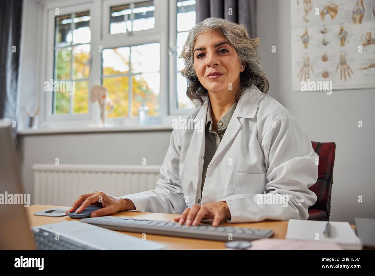 Ritratto del GP femminile maturo indossando cappotto bianco alla scrivania in ufficio medico Foto Stock