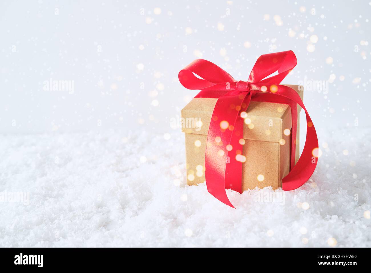 Confezione regalo artigianale con un arco rosso nella neve. Sfondo bianco e luci festive. Foto Stock