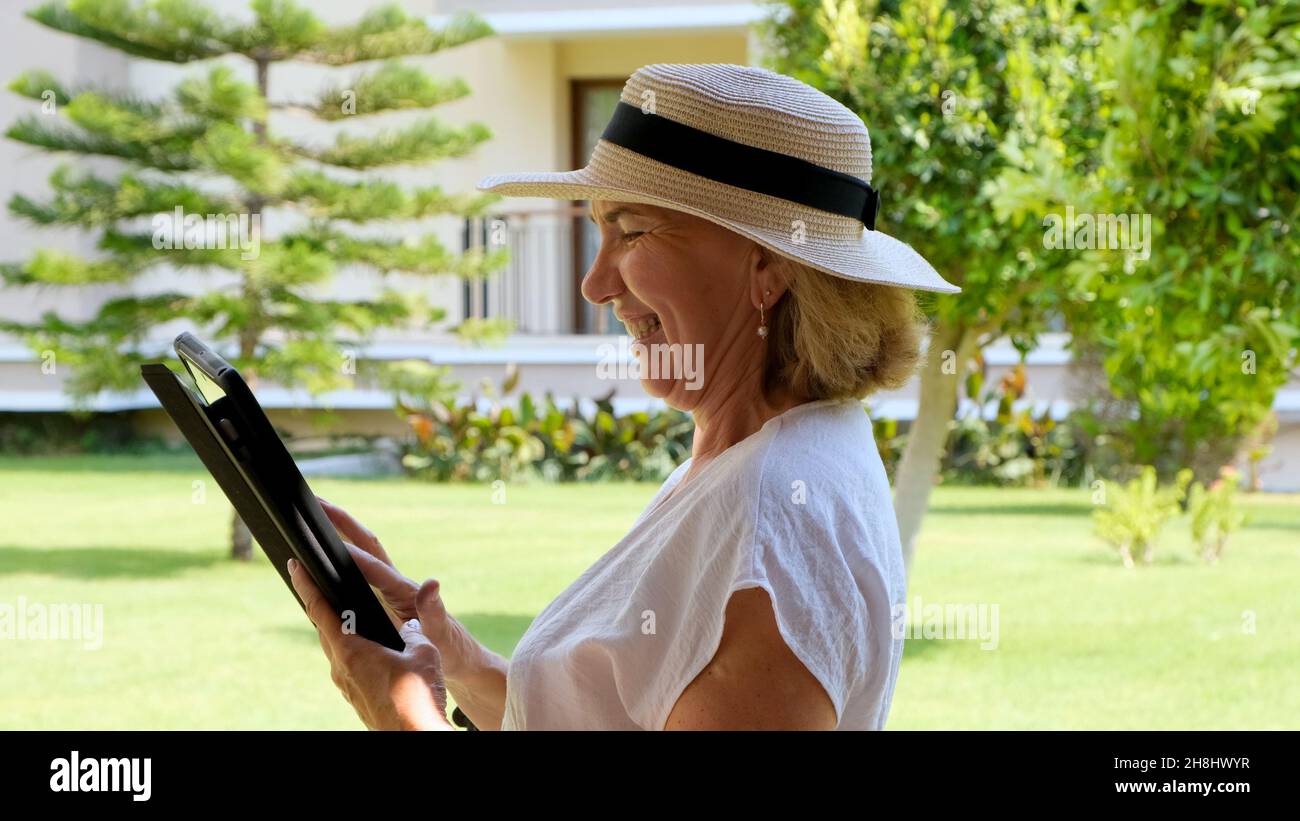 Donna anziana donna d'affari bionda in cappello usa un tablet pc, un tablet digitale per lavoro d'affari o per studio nel suo giardino verde. Donna di età compresa tra 50 e 55 anni. Il Foto Stock