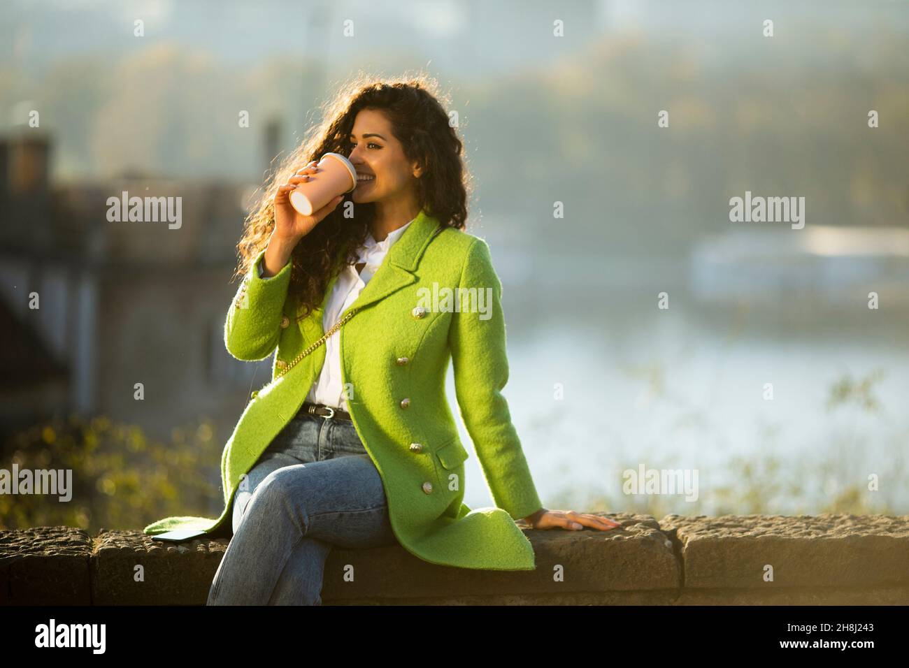 Bella giovane donna capelli ricci godendo il sole autunnale mentre si siede accanto al fiume e bere caffè takeaway Foto Stock