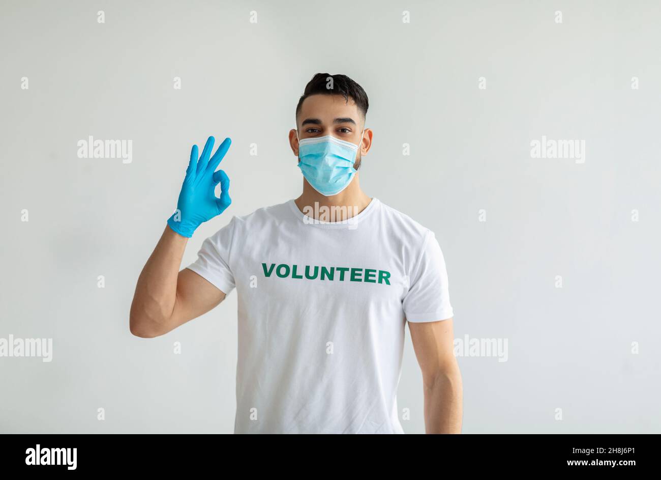 Volontariato libero, beneficenza grazia concetto. Volontario maschile in maschera medica e guanti mostrando OK gesto, parete di luce Foto Stock