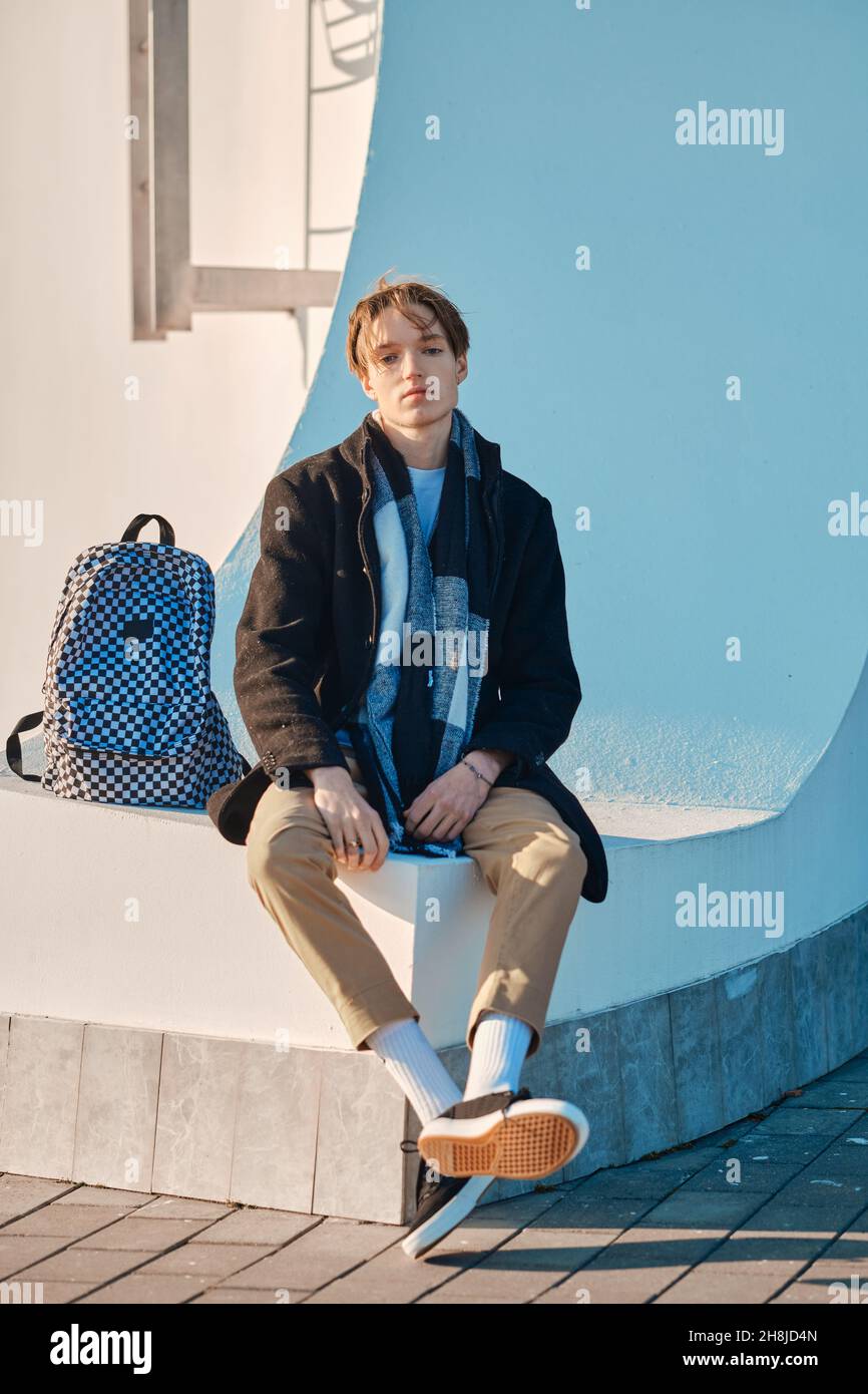 Giovane studente con zaino in vecchio cappotto di lana, sciarpa e pantaloni corti si siede all'aperto Foto Stock