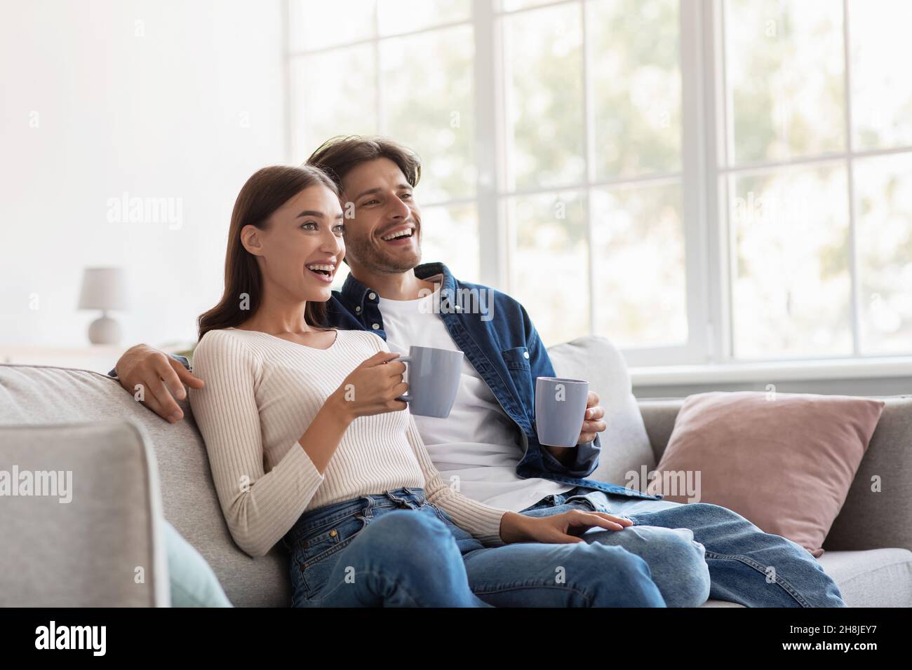 Sorridente giovane europeo uomo e donna rilassato con tazze di bevanda calda godere di tempo libero e guardare il film Foto Stock