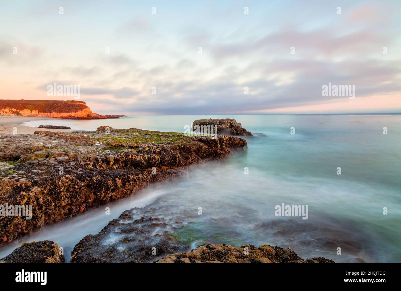 L'immagine cattura una tranquilla scena costiera al tramonto, caratterizzata da rocce scoscese ricoperte di muschio che si proiettano nell'oceano turchese e calmo. Foto Stock