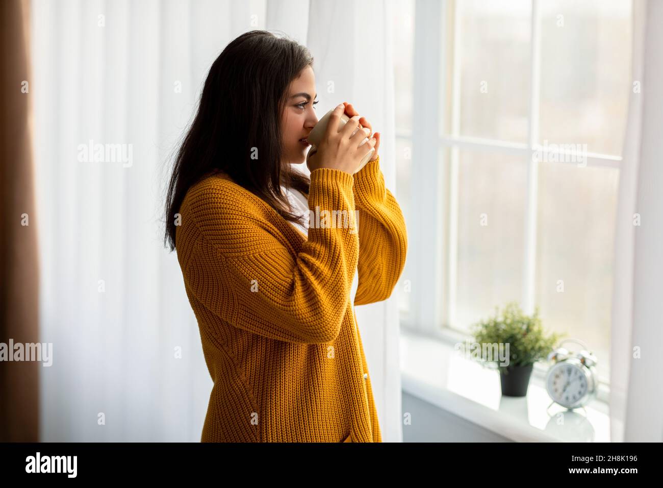 Giovane ragazza araba bevendo bevande calde e guardando fuori dalla finestra, chillando a casa la Domenica mattina, spazio copia Foto Stock