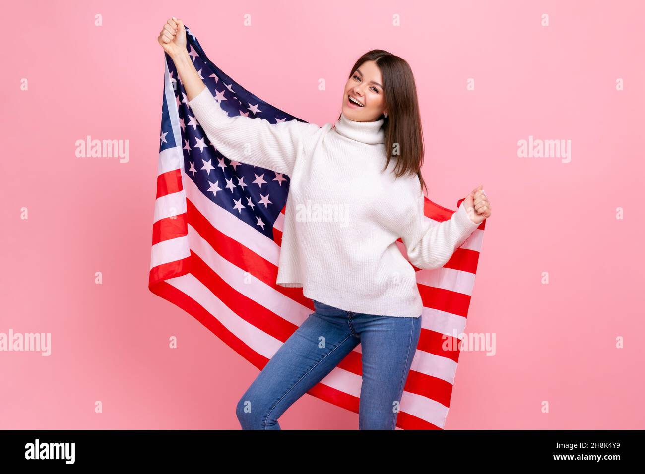 Ritratto di bella donna braccia sollevate, tenendo bandiera americana, festeggiamenti nazionali, ballando, indossando bianco casual stile maglione. Studio interno girato isolato su sfondo rosa. Foto Stock