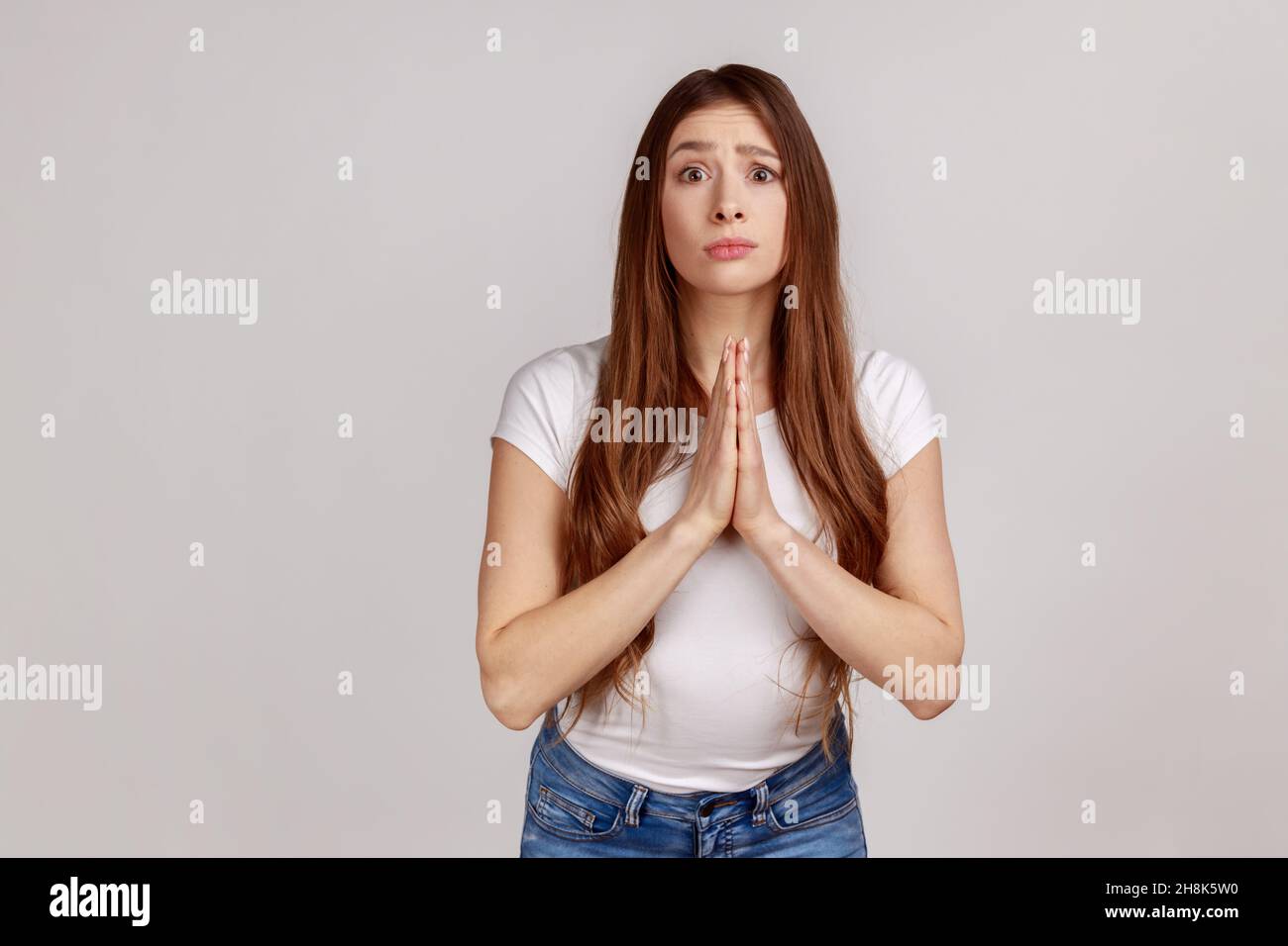 Per favore, ho bisogno. Giovane adulta che tiene le mani in preghiera e chiede aiuto con implorazione implorazione, piena di speranza, indossando una T-shirt bianca. Studio interno girato isolato su sfondo grigio. Foto Stock