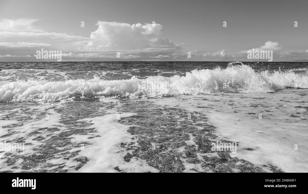 Schiuma splendente sulla spiaggia di ciottoli con onde tempestose e nubi epiche sulla costa dell'isola di Lefkada in Grecia. Viaggio nel Mar Ionio in scala di grigi Foto Stock